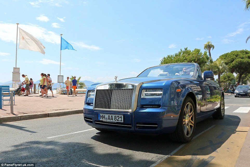 A man drives his large blue convertible down a seafront road in Cannes while walkers stop and stare at the vehicle 